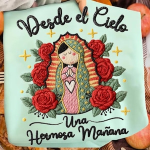 May include: A light teal embroidered pillow with the text "Desde el Cielo" and "Una Hermosa Mañana". The design features a religious figure with a heart, surrounded by red roses and gold crosses.