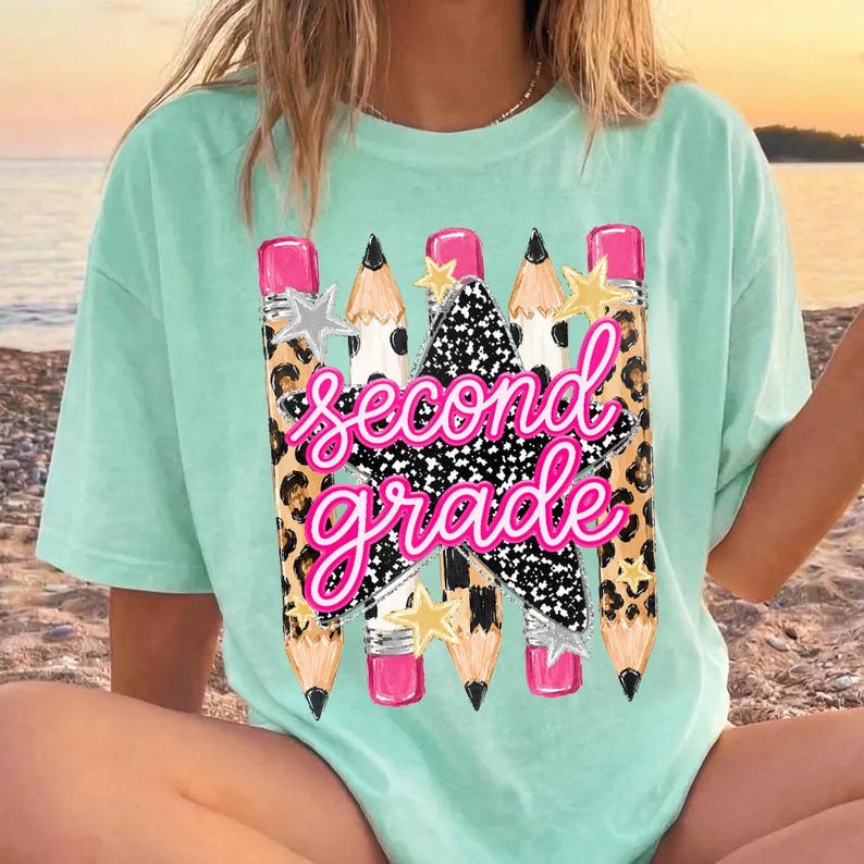 May include: A light mint green t-shirt featuring a graphic print of four pencils with leopard print patterns and pink erasers. A black and white speckled star is centered behind the bright pink cursive text that reads second grade.
