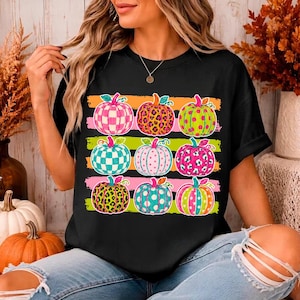 May include: A black short-sleeve t-shirt featuring a graphic print of nine pumpkins arranged in three rows. The pumpkins display various patterns including checkered, leopard print, and polka dots in vibrant shades of pink, green, and orange.