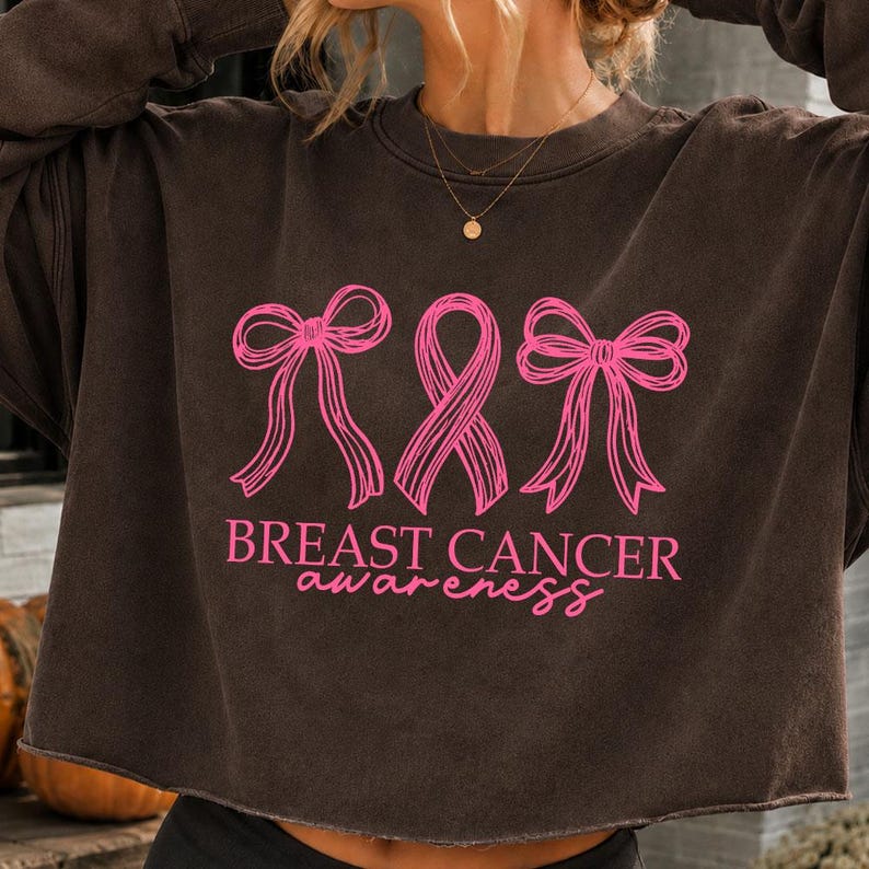 May include: A dark brown long-sleeve sweatshirt featuring a pink graphic of two bows flanking a breast cancer awareness ribbon. The text "BREAST CANCER awareness" is printed in pink below the graphic. The sweatshirt has a raw, unfinished hem.