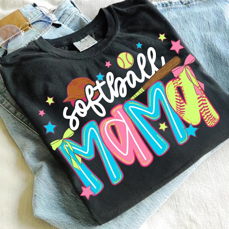 May include: Black t-shirt with the words "Softball MAMA" in a colorful design. The design includes a softball, bat, and glove. The shirt is folded on top of a pair of blue jeans.