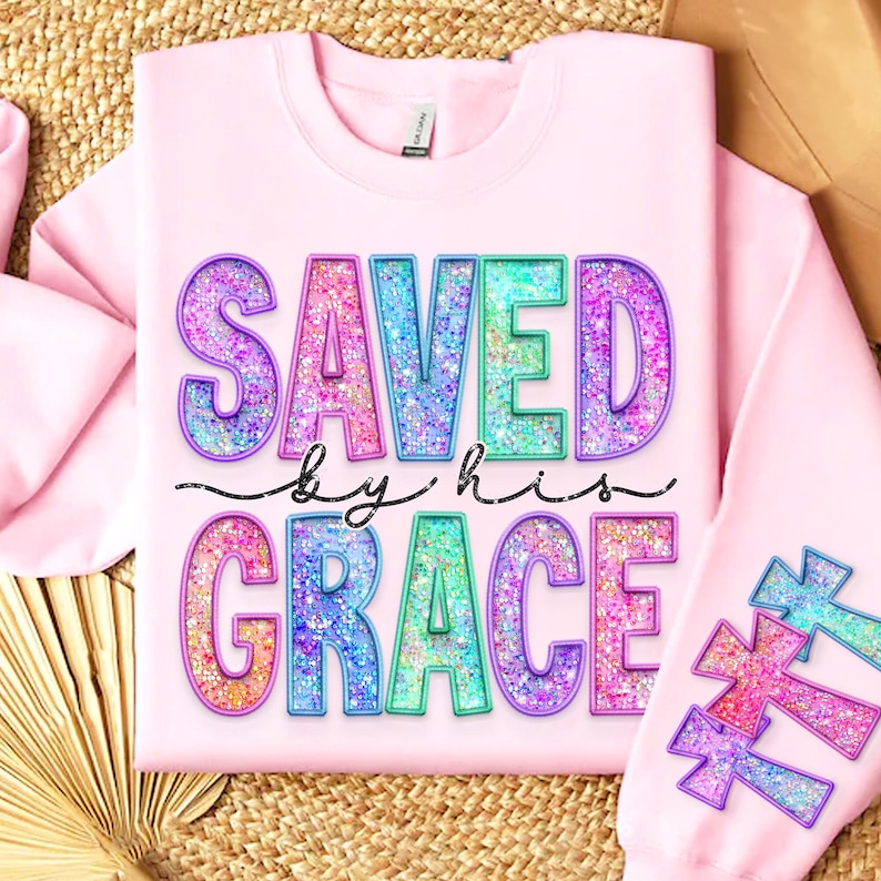 May include: Light pink sweatshirt with the words "SAVED by his GRACE" in large, glittery, colorful letters. The sleeve features glittery crosses in shades of pink, blue, and green. The sweatshirt is made of a soft material.