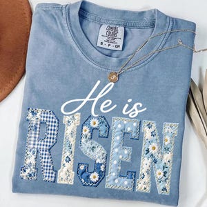 May include: A pale blue Comfort Colors t-shirt with the words "He is RISEN" in white script and patchwork letters. The letters are decorated with blue floral and gingham patterns. A gold necklace with a pendant rests on the shirt.