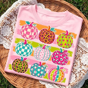 May include: A light pink t-shirt featuring a graphic print of nine pumpkins arranged in three rows. The pumpkins display various patterns including checkered, leopard print, and polka dots in vibrant colors like orange, green, and pink.