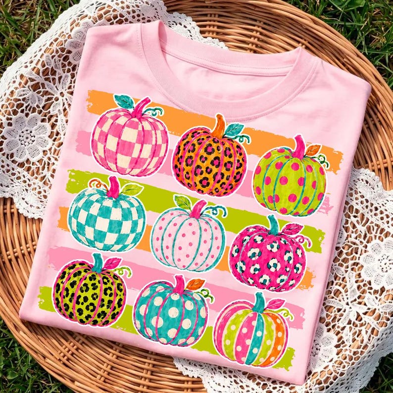 May include: A light pink t-shirt featuring a graphic print of nine pumpkins arranged in three rows. The pumpkins display various patterns including checkered, leopard print, and polka dots in vibrant colors like orange, green, and pink.