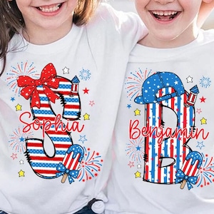 May include: Two white t-shirts with patriotic designs. One shirt features the name "Sophia" with a large "3" in red, white, and blue, a bow, fireworks, and an ice pop. The other shirt has the name "Benjamin" with a "B" and a baseball cap.