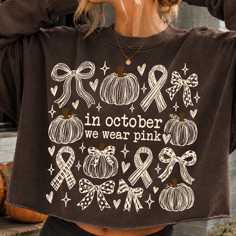 May include: A dark brown long-sleeve sweatshirt featuring a white graphic print of pumpkins, ribbons, and bows. The text in the center reads, "in october we wear pink," surrounded by small heart and star icons, with a crew neckline and raw hem.