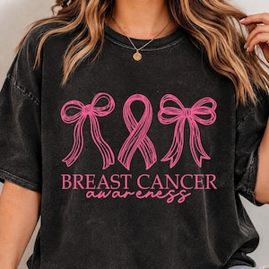 May include: A black short-sleeved t-shirt with a graphic print in pink. The design features two bows flanking a central awareness ribbon. Below the graphic, the text reads BREAST CANCER awareness in a combination of block and script fonts.