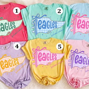 以下が含まれることがあります： パステルカラーや鮮やかな色の半袖クルーネックTシャツ6枚。それぞれにギンガムチェック柄のペナントフラッグのグラフィックが描かれており、「EAGLES」と「School name」の文字が筆記体でプリントされています。1から6までの番号が振られています。