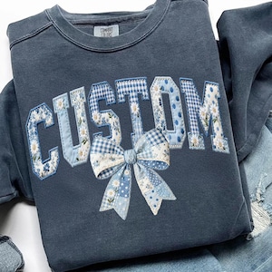 May include: A navy blue sweatshirt with the word "CUSTOM" appliquéd in a patchwork design. The letters feature floral, polka dot, and gingham patterns in shades of blue and white. A matching bow is centered below the letters. The tag reads "COMFORT COLORS".
