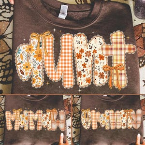 May include: A brown sweatshirt with the word "MOM" in a patchwork design. The letters feature floral, gingham, and plaid patterns in orange, yellow, and white, with decorative bows. The sweatshirt is a fall-themed apparel item.