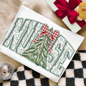 Op de afbeelding: Wit kussen met het woord "MERRY" in een groen gestikt lettertype. De letters zijn gevuld met een bloemenpatroon en een kerstboomontwerp. Een rood-witte geruite strik staat op de boom. Een geschenkdoos met een rode strik en een peperkoeken man staan op de achtergrond.