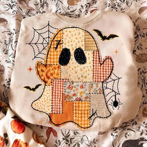 May include: A cream-colored sweatshirt featuring a large central graphic of a ghost composed of various patchwork patterns in shades of orange, yellow, and white. The design includes black spiderwebs, bats, and a spider, with two solid black oval eyes.
