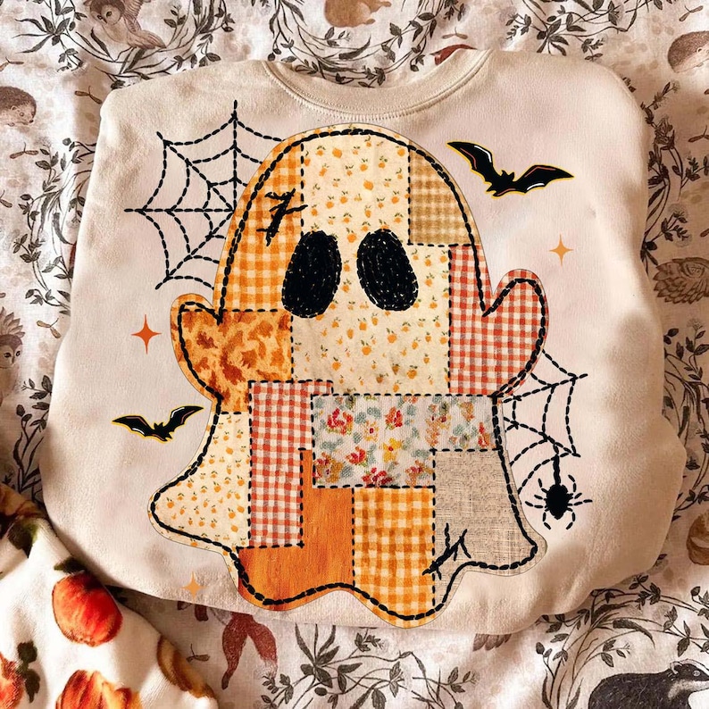May include: A cream-colored sweatshirt featuring a large central graphic of a ghost composed of various patchwork patterns in shades of orange, yellow, and white. The design includes black spiderwebs, bats, and a spider, with two solid black oval eyes.