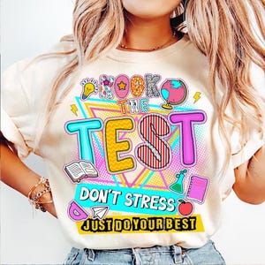 May include: A white t-shirt with a colorful graphic design that says "Rock the Test Don't Stress Just Do Your Best". The graphic includes a lightbulb, a star, a globe, a book, a test tube, a notebook, and an apple.