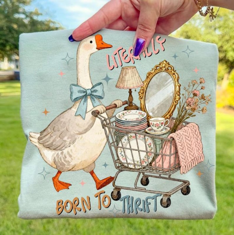 May include: A light blue graphic tee featuring a goose pushing a shopping cart filled with vintage items. The goose wears a blue bow tie and the text reads "Literally Born to Thrift". The cart contains plates, a mirror, a lamp, and a teacup.
