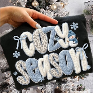 Pu&ograve; includere: T-shirt nero con le parole "COZY SEASON" in stile lavorato a maglia, con accenti blu e bianchi, dettagli animalier, fiocchi di neve e una grafica a pupazzo di neve. Un design festivo a tema invernale.