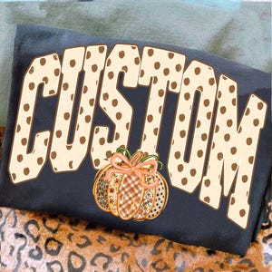 Pode incluir: Uma t-shirt cinzento-escura com a palavra CUSTOM em letras grandes de bloco cor de creme com pintas castanhas. Abaixo do texto encontra-se um gráfico de abóbora com um laço laranja claro, decorado com padrões xadrez, florais e de leopardo.