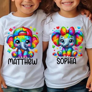 以下が含まれることがあります： カラフルなパズルピースの象のデザインが施された白いTシャツ2枚。左側のシャツには「マシュー」と書いてあり、右側のシャツには「ソフィア」と書いてあります。