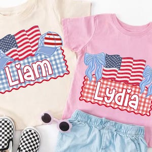 Puede incluir: Dos camisetas infantiles, una crema y otra rosa, con dise&ntilde;os patri&oacute;ticos. La camiseta crema presenta banderas, gorras y el nombre "Liam". La camiseta rosa tiene banderas, lazos y el nombre "Lydia".