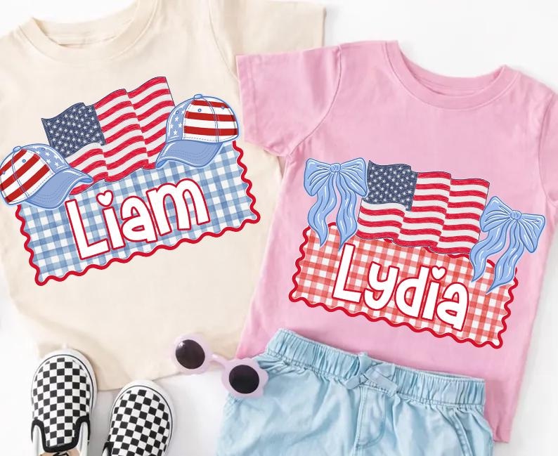 Puede incluir: Dos camisetas infantiles, una crema y otra rosa, con dise&ntilde;os patri&oacute;ticos. La camiseta crema presenta banderas, gorras y el nombre "Liam". La camiseta rosa tiene banderas, lazos y el nombre "Lydia".