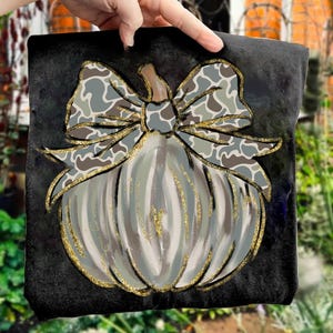 May include: A black square canvas features a painted pumpkin with a bow. The pumpkin is light gray with vertical stripes and gold glitter accents. The bow has a camouflage pattern in shades of gray and brown.