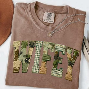 Op de afbeelding: Een bruin Comfort Colors t-shirt met het woord "WIFEY" op de voorkant. De letters zijn een mix van camouflage- en ruitpatronen. Een zilveren ketting met een kleine hanger ligt over het shirt.