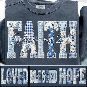 May include: A navy blue sweatshirt with the word "FAITH" in a patchwork design, featuring floral, gingham, and polka dot patterns in shades of blue and white. The text "Ephesians 2:8" is below the word "FAITH". Additional designs include "LOVED", "BLESSED", and "HOPE".