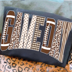 May include: A dark gray t-shirt featuring the text GAME DAY in large block letters. The letters are filled with various patterns including brown footballs with white laces, leopard print, brown polka dots, brown plaid, and brown vertical stripes.