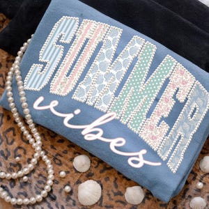 May include: A blue sweatshirt with the words "SUMMER vibes" in a decorative font. The letters are filled with various patterns, including stripes, shells, and floral designs. A pearl necklace and seashells are arranged around the sweatshirt.