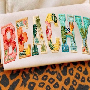 Puede incluir: Tela blanca bordada con la palabra "BEACHY" en letras coloridas con motivos florales. Las letras están delineadas en dorado y presentan varios diseños de flores y hojas tropicales, incluyendo hibiscos y aves del paraíso.
