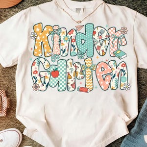 May include: White t-shirt with the word "Kindergarten" in colorful, patterned letters. The letters feature floral, polka dot, and checkered designs, with school-themed illustrations like scissors and pencils.