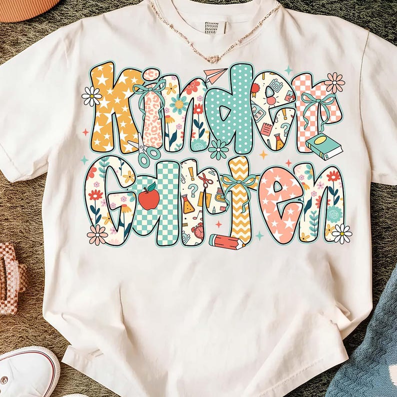 May include: White t-shirt with the word "Kindergarten" in colorful, patterned letters. The letters feature floral, polka dot, and checkered designs, with school-themed illustrations like scissors and pencils.