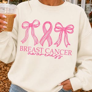 May include: A cream-colored crewneck sweatshirt featuring a pink graphic design of two bows flanking a breast cancer awareness ribbon. Below the graphic, the text reads BREAST CANCER awareness in a combination of block and script lettering.