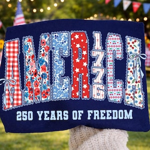 May include: Navy blue sweatshirt with the word "AMERICA" in a patriotic design. The letters are filled with patterns like gingham, floral, and stars. The text "250 YEARS OF FREEDOM" is printed below the word AMERICA.
