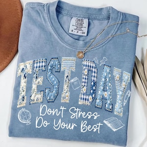 May include: A light blue Comfort Colors t-shirt with the words "TEST DAY" in a patchwork design. The shirt also features the text "Don't Stress Do Your Best" and small illustrations. A gold necklace is draped across the shirt.