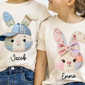 May include: Two off-white t-shirts featuring bunny face appliqués. One bunny wears a blue and white plaid cap, the other has a floral bow. The name "Jacob" is below one bunny, and "Emma" is below the other.