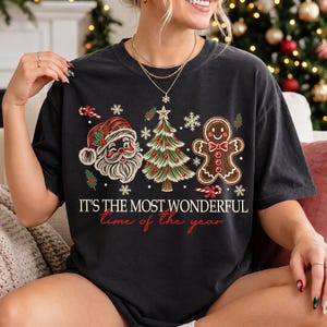 K&ouml;nnte beinhalten: Ein schwarzes Kurzarm-T-Shirt mit einem Grafikdruck von Weihnachtsmann, einem geschm&uuml;ckten Weihnachtsbaum und einem Lebkuchenmann. Der Text lautet "IT'S THE MOST WONDERFUL time of the year" in wei&szlig;er und roter Schrift, umgeben von Schneeflocken.