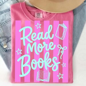 Peut inclure: T-shirt rose avec des rayures verticales roses et l'inscription "Read More Books" en bleu clair. Le motif comprend des &eacute;toiles et des illustrations de livres. Le t-shirt est pli&eacute; sur un jean bleu.