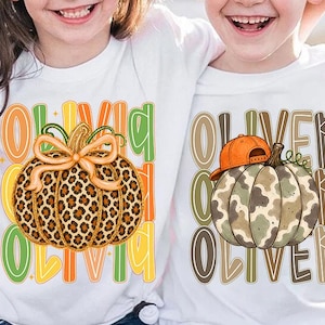 May include: Two white t-shirts featuring pumpkin graphics. The left shirt displays a leopard print pumpkin with a bow and the name OLIVIA. The right shirt shows a camouflage pumpkin wearing an orange baseball cap with the name OLIVER.