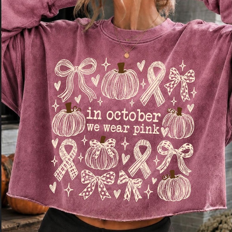 May include: A mauve long-sleeve sweatshirt with a raw hem featuring a white graphic print of pumpkins, bows, and awareness ribbons. The text in the center reads, in october we wear pink, surrounded by small heart and star shapes across the front.