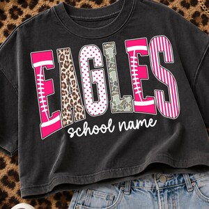 May include: A charcoal gray cropped t-shirt featuring the word EAGLES in large, multi-patterned letters including pink football laces, leopard print, pink polka dots, camouflage, and pink stripes, with the white cursive text school name below.