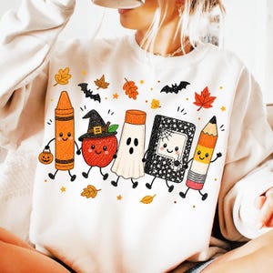 May include: A white crewneck sweatshirt featuring a colorful graphic print of five anthropomorphic school supplies and a red apple. The characters include an orange crayon, a red apple wearing a witch hat, a white ghost, a black notebook, and a yellow pencil.