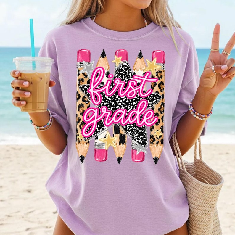 May include: A light purple t-shirt featuring a graphic print of four pencils with leopard print patterns and pink erasers. A large black and white speckled star is centered with the text "first grade" in bright pink cursive lettering.