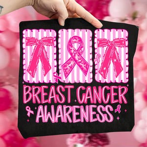 May include: Black tote bag with pink ribbon graphics and the words "Breast Cancer Awareness" in pink. The design includes pink bows and a pink ribbon symbol. The bag is held by a hand.
