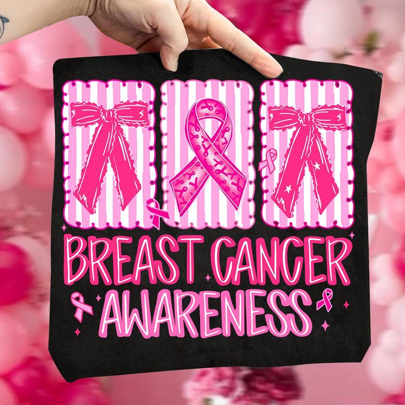 May include: Black tote bag with pink ribbon graphics and the words "Breast Cancer Awareness" in pink. The design includes pink bows and a pink ribbon symbol. The bag is held by a hand.