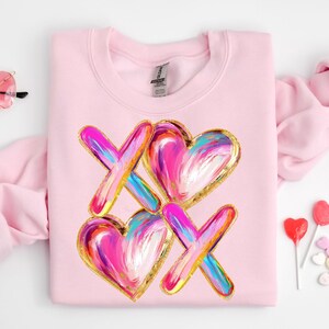 Puede incluir: Sudadera rosa con un dise&ntilde;o gr&aacute;fico de dos corazones y dos X en un estilo abstracto y colorido con detalles dorados.