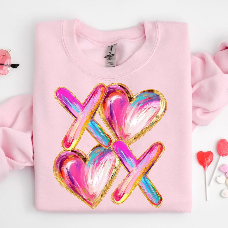 Puede incluir: Sudadera rosa con un dise&ntilde;o gr&aacute;fico de dos corazones y dos X en un estilo abstracto y colorido con detalles dorados.