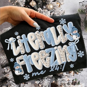 May include: Black sweatshirt with the text "Literally Freezing me 24:7" in a knitted style with blue and white accents. Features snowflakes and a snowman graphic. A festive winter-themed apparel item.