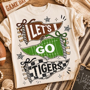 May include: A cream-colored t-shirt featuring a graphic design of three pennants. The top brown pennant says LET'S, the middle green football field pennant says GO, and the bottom checkered pennant says TIGERS. The design includes stars and accents.
